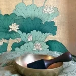 A metal singing bowl with a wooden mallet resting on a patterned cushion in front of a lotus-flower backdrop